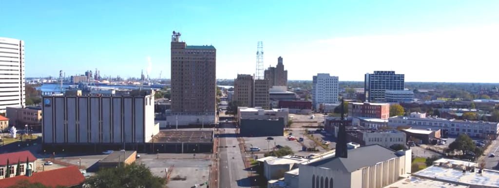 A wide view of downtown Beaumont's low skyline under a clear sky, with mid-rise buildings in the foreground and refinery structures and port cranes visible along the horizon to the left.