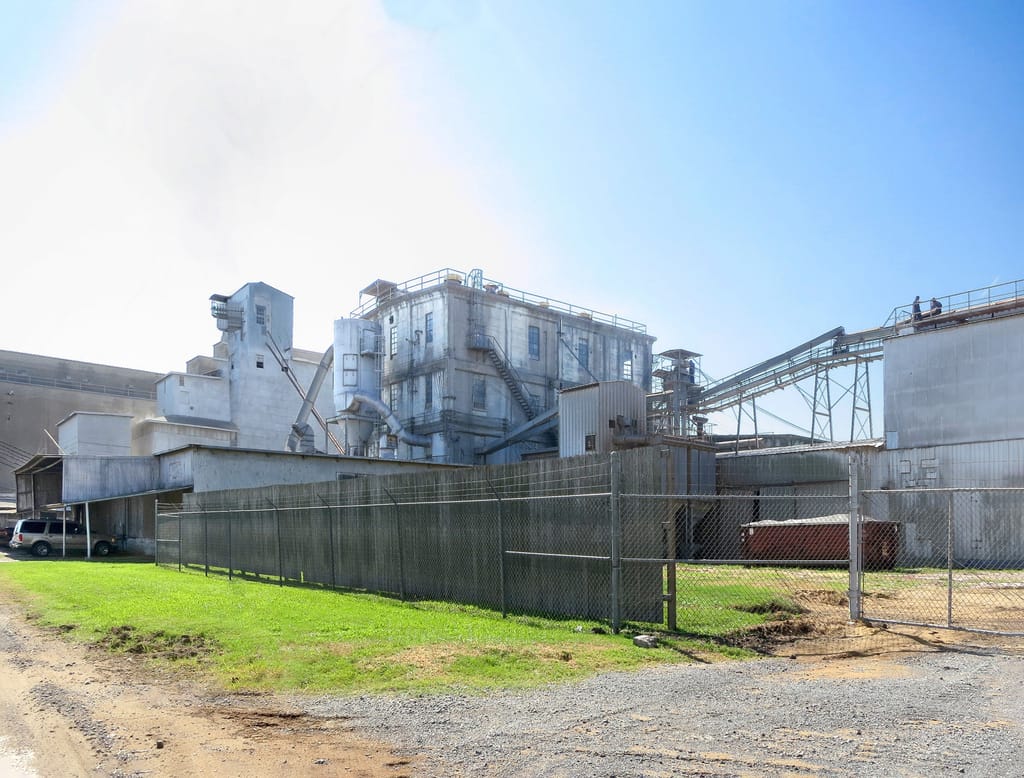 A large early-twentieth-century concrete rice mill in Beaumont: tall silos and processing structures behind a chain-link fence, with conveyors and a grassy verge in the foreground under a bright sky.