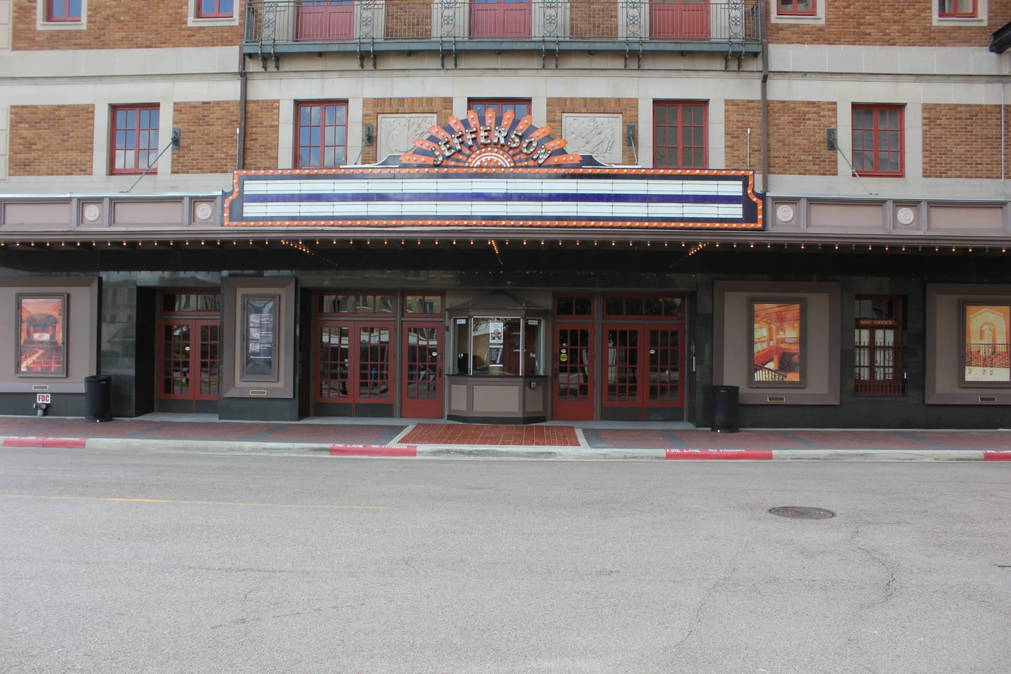 The Jefferson Theatre facade in downtown Beaumont: tan brick, red-trimmed windows and doors, and an illuminated sunburst marquee spelling JEFFERSON above a street-level ticket kiosk marked Box Office.