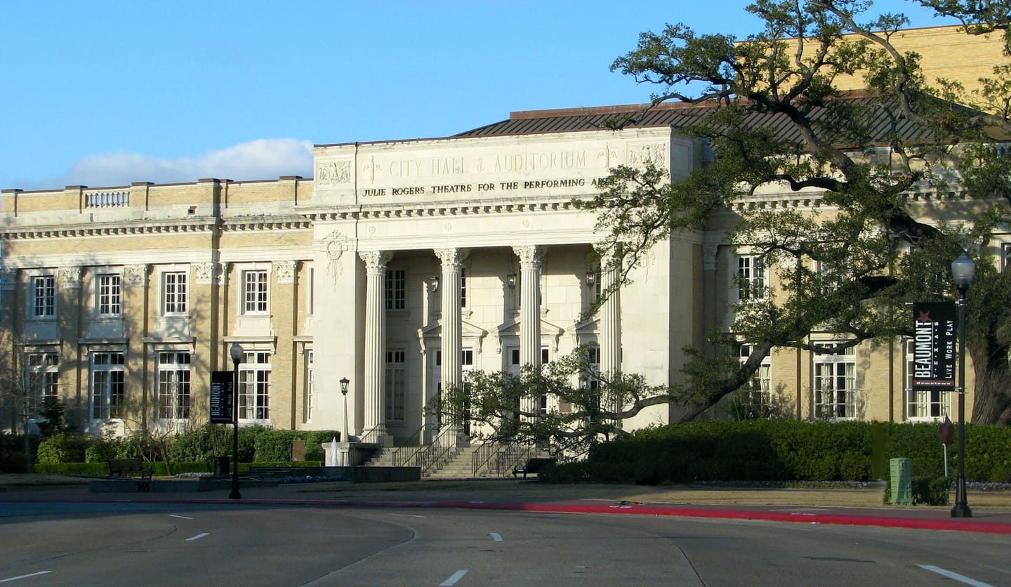 The Julie Rogers Theatre in Beaumont, a 1928 neoclassical building with Corinthian columns and the carved inscription CITY HALL AND AUDITORIUM above JULIE ROGERS THEATRE FOR THE PERFORMING ARTS, framed by a live oak.