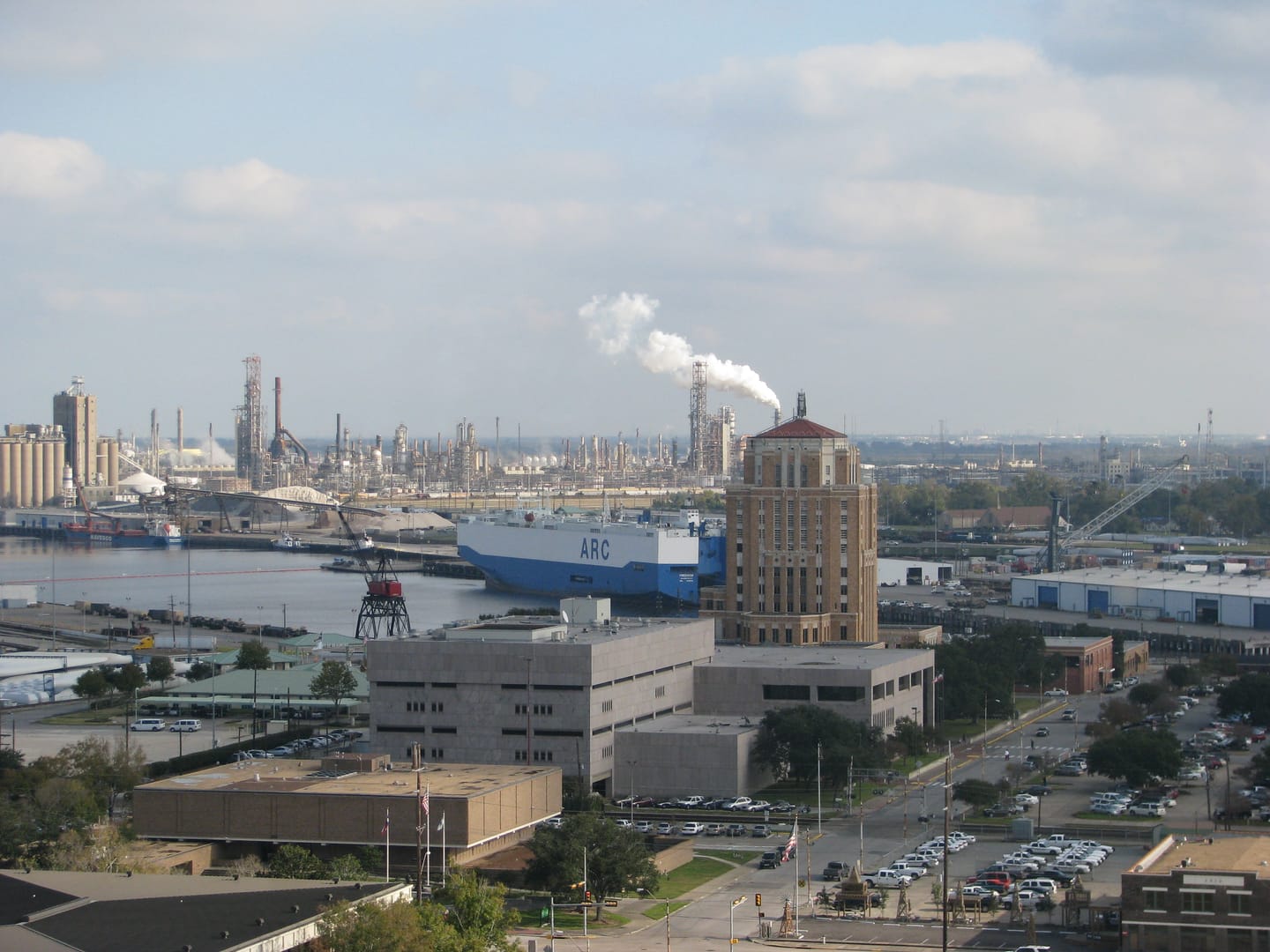 The Port of Beaumont from above: a blue and white roll-on/roll-off car carrier docked along the Neches River, dockside cranes and cargo, refinery towers with a steam plume in the distance, and the Jefferson County courthouse tower rising over downtown.