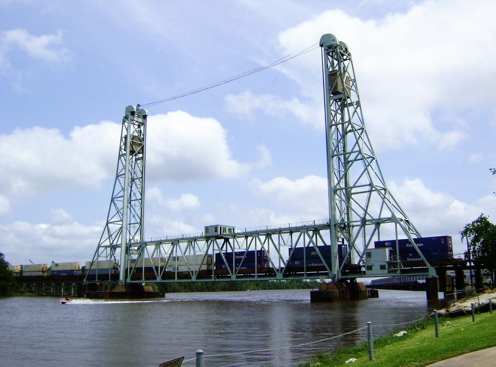 A steel vertical-lift truss railroad bridge over the Neches River seen from Riverfront Park in Beaumont, with a container freight train crossing the raised span between its two lift towers and a park railing in the foreground.
