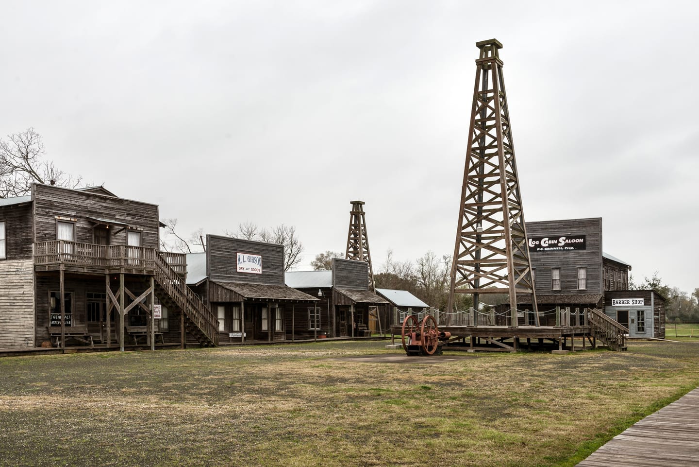 A modern reconstruction of the Spindletop-Gladys City boomtown near Beaumont: weathered wooden false-front buildings labeled A. L. Gibson Dry Goods, Log Cabin Saloon, and Barber Shop, with tall wooden oil derricks rising behind them under an overcast sky.